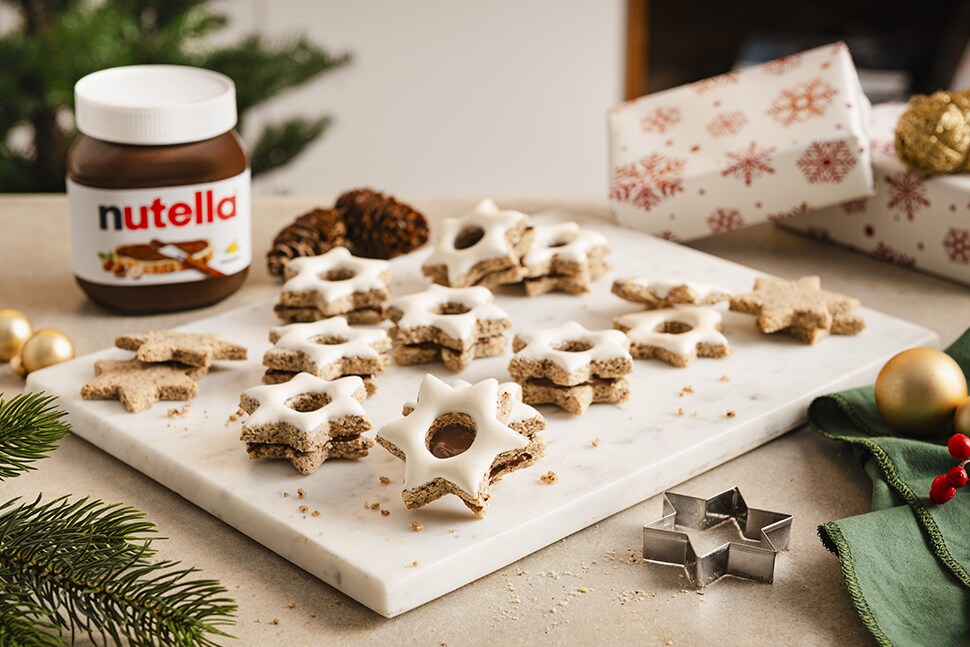 Mehrere frisch gebackene und glasierte Zimtsterne liegen auf einer weißen Marmorplatte. Neben der Marmorplatte steht ein Glas nutella®.