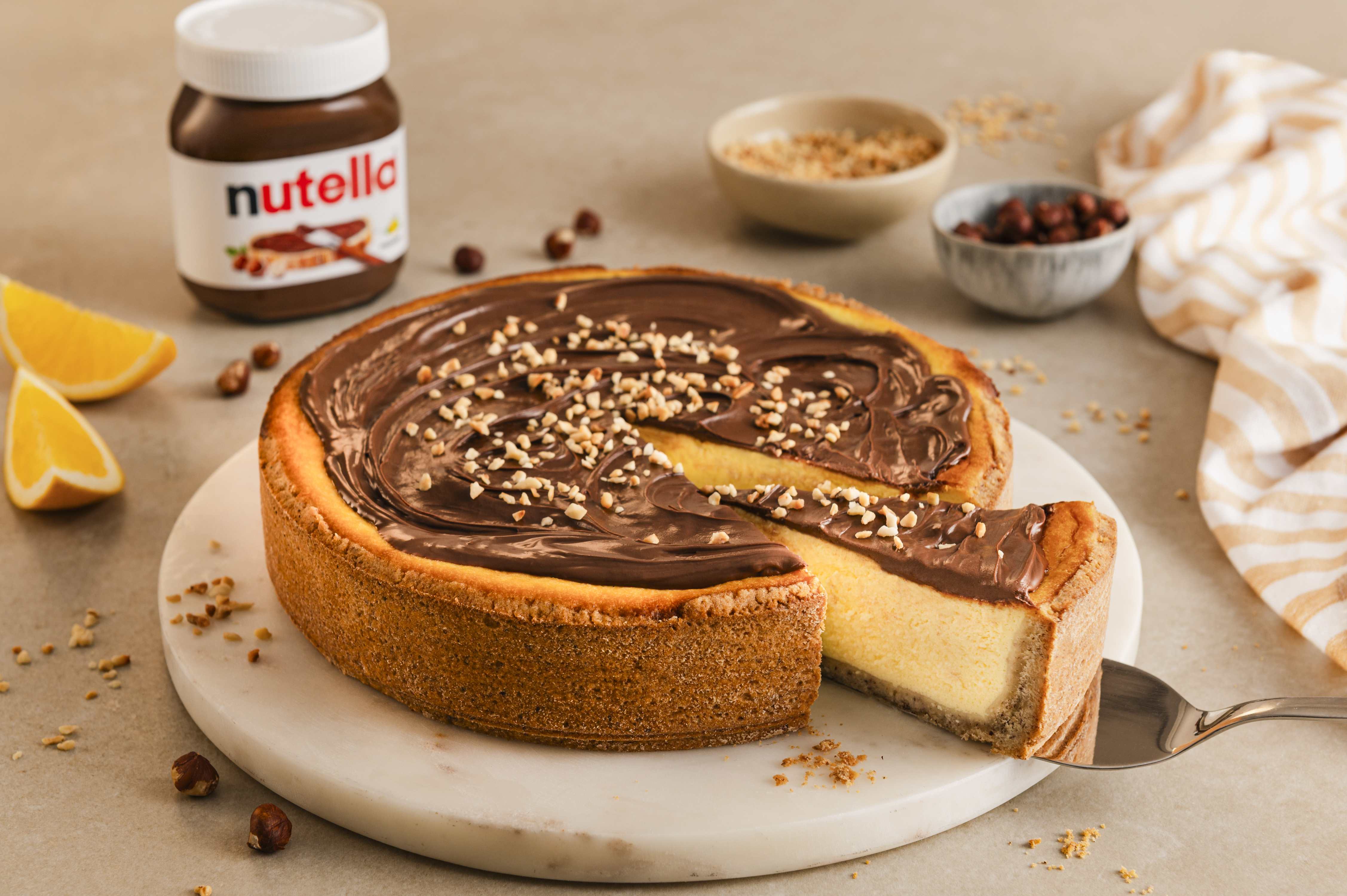 Gebackener Käsekuchen mit nutella® und Nüssen, ein Stück wird serviert. Dazu gibt es nutella®, Orangen und Nüsse.