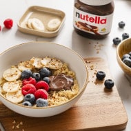 Frühstücks-Bowl mit Quinoa, Banane, Beeren und nutella, umgeben von frischen Zutaten