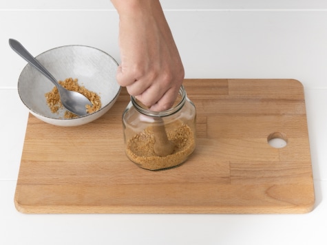 Keksbrösel werden in einem Glas mit einem Stößel angedrückt, daneben eine Schale mit weiteren Bröseln