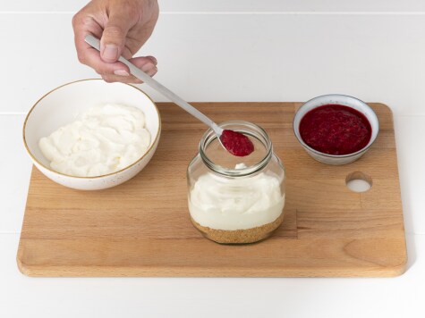 Himbeerpüree wird mit einem Löffel auf eine Schicht Cheesecake-Creme im Glas gegeben, daneben Zutaten in Schalen