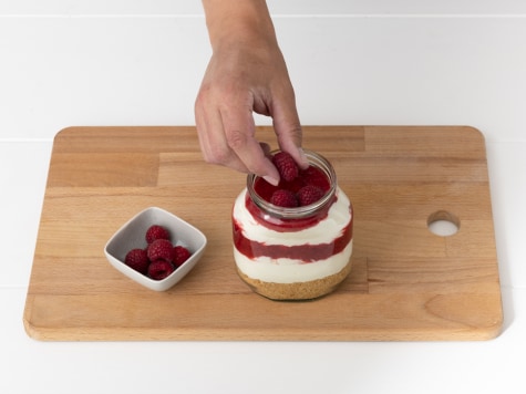 Frische Himbeeren werden mit der Hand auf das fertige Dessert im Glas gegeben, daneben eine kleine Schale mit Himbeeren