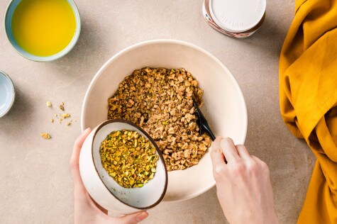 Overhead-Aufnahme von Händen, die gehackte Pistazien in eine Schüssel mit gemischten Nüssen geben, mit einem schwarzen Spatel, einer Schüssel mit gelber Flüssigkeit und einem Glas Nutella in der Nähe.