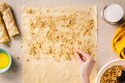 Overhead-Aufnahme einer Person, die gehackte Nüsse und Pistazien auf Filoteig streut, mit gerolltem Baklava, geschmolzener Butter, Nutella und einer Schüssel mit Nussmischung in der Nähe.