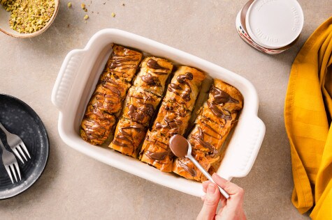Overhead-Aufnahme von Nutella-Baklava in einer weißen Auflaufform, beträufelt mit Nutella, mit einem Löffel, Pistazien, Gabeln, einem Nutella-Glas und einem gelben Tuch in der Nähe