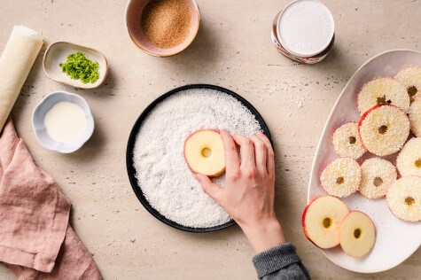 Apfelscheibe wird in Kokosraspeln gewendet, daneben Zutaten wie Blätterteig, Limettenabrieb und brauner Zucker