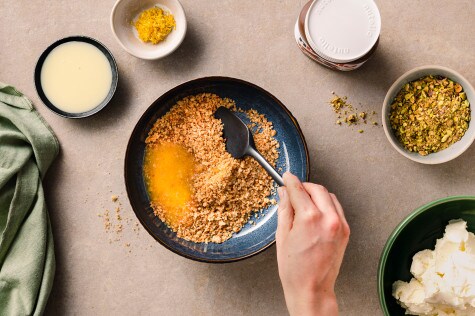 Butter wird in einer Schüssel mit zerkleinerten Haferkeksen vermengt, daneben Zutaten wie Zitronenabrieb, Kondensmilch und gehackte Pistazien