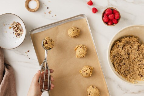 Ein Backblech mit Backpapier ist mit vier Teigkugeln für die Cookies belegt. Dabei ist die Verwendung eines silbernen Eisportionierers zu erkennen.