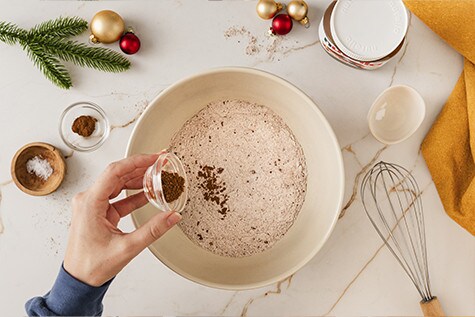 In einer Rührschüssel wird eine Kakaomehlmischung mit aromatischen Lebkuchengewürzen verfeinert. Auf dem Tisch liegen weitere weihnachtliche Zutaten.