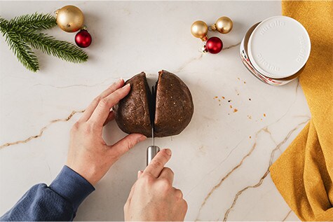Ein Lebkuchenteig ist zu einer Kugel geformt und liegt auf einer Arbeitsfläche bereit. Die Kugel wird mit einem Messer in zwei Hälften zerteilt.