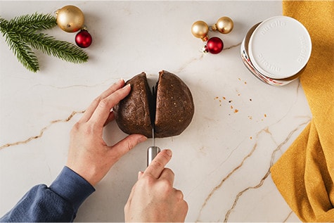 Ein Lebkuchenteig ist zu einer Kugel geformt und liegt auf einer Arbeitsfläche bereit. Die Kugel wird mit einem Messer in zwei Hälften zerteilt.