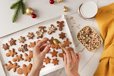 Frisch gebackene Weihnachts-Lebkuchen-Kekse in Stern- und Figurenform liegen auf einem Blech. Mit einem Löffel wird nutella® auf den Keks gestrichen.