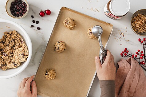 Ein Blech mit Backpapier liegt in der Mitte des Tisches. Auf diesem werden mit einem Eisportionierer Kugeln aus Haferflocken-Cranberry-Teig gesetzt.