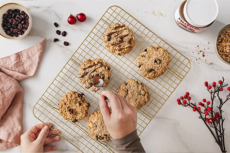 Frisch gebackene Haferflockenkekse auf einem goldenen Gitter. Eine Hand verteilt mit einem Löffel hauchzarte Streifen nutella® auf die Kekse.