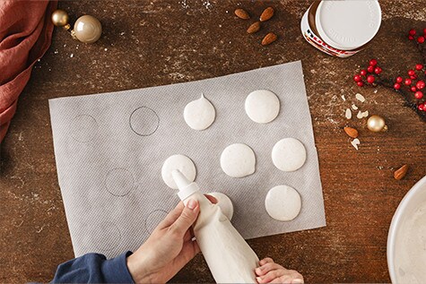 Mandelmasse in einem Spritzbeutel wird kreisförmig auf ein Stück Backpapier gepresst. Ein Glas nutella® und weihnachtliche Deko sind auf dem Tisch platziert.