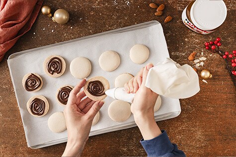 Eine Hand drückt mit dem Spritzbeutel die nutella® Füllung spiralförmig in die Macaron-Halbkugeln hinein, während die andere die Macaron-Hälfte hält.