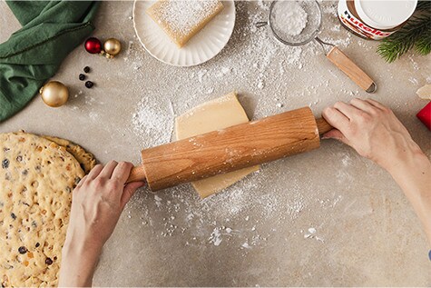 Ein Stück Marzipan wird auf einer bemehlten Arbeitsfläche mit dem Nudelholz ausgerollt. Daneben liegt der ausgerollte Hefeteig mit Rosinen