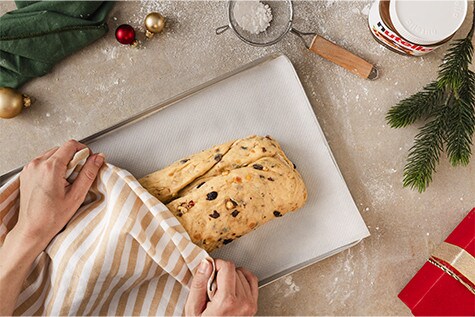 Ein gefüllter Stollenteig liegt auf einem Backblech mit Backpapier. Mit einem gestreiften Küchentuch wird er zum Ruhen abgedeckt.