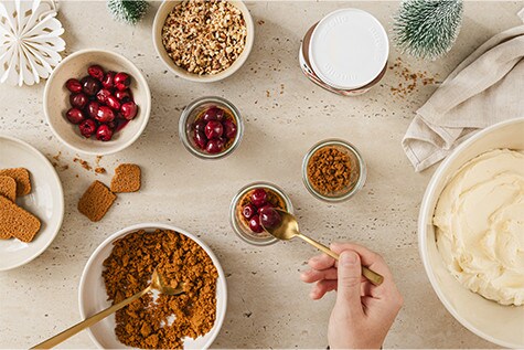 Kleine Dessertgläser werden mit einer Schicht Spekulatius-Bröseln gefüllt. Darauf platziert sind frische rote Kirschen.