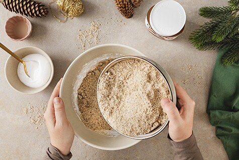 Eine aufgeschlagene Eiweißmasse befindet sich in einer großen Schüssel, die mit gemahlenen Haselnüssen bestreut wird.