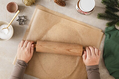 Auf einem Tisch wird ein Teig zwischen zwei Blättern Backpapier mit einem hölzernen Nudelholz ausgerollt.