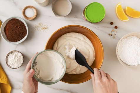 Draufsicht auf Hände, die Frischkäse in eine Schüssel geben; weitere Backzutaten und ein Glas nutella® Plant-Based stehen daneben