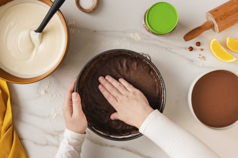 Draufsicht auf Hände, die Schokoteig in eine Backform drücken; Schüsseln mit Sahnecreme und ein Glas nutella® Plant-Based daneben