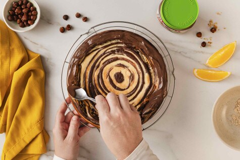 Hände verstreichen Schokoladenguss auf einem Zebrakuchen; Haselnüsse und ein Glas nutella® Plant-Based daneben