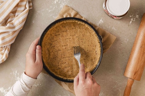 Draufsicht auf Hände, die mit einer Gabel Löcher in einen Teigboden stechen; nutella® und ein Nudelholz liegen daneben.