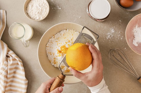 Draufsicht auf Hände, die Orangenschale in eine Schüssel mit Backzutaten reiben; daneben ein Schneebesen, Milch und ein Glas nutella®.