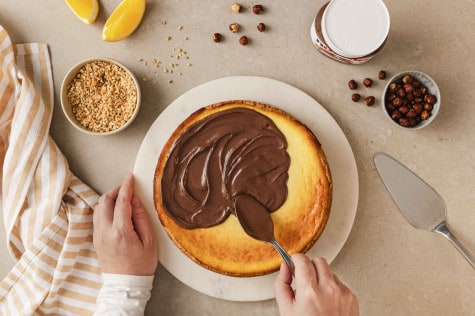 Draufsicht auf Hände, die nutella® auf Käsekuchen verteilen. Nüsse, Orangen und ein nutella®-Glas stehen daneben.