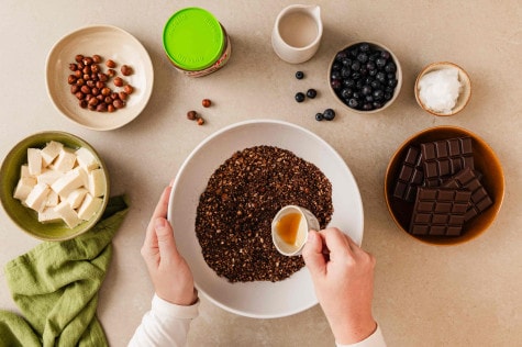 Hände gießen Ahornsirup über fein gemahlene Haselnüsse in Schüssel; Nüsse, Tofu, Beeren, Schokolade, Glas nutella® Plant-Based daneben.