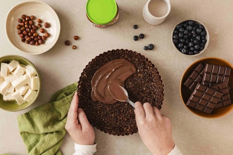 Hände verstreichen nutella® Plant-Based auf Nusskuchenboden; Nüsse, Tofu, Blaubeeren, Schokolade daneben. 