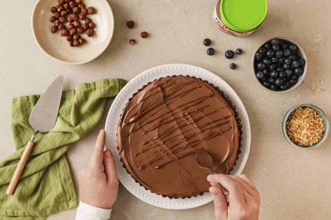 Draufsicht auf Hände, die Schokoladenguss auf einen Kuchen träufeln; Nüsse, Blaubeeren und ein Glas nutella® Plant-Based stehen daneben