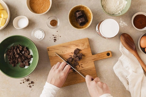 Draufsicht auf Hände, die Schokolade auf einem Holzbrett hacken, daneben Backzutaten und ein nutella®-Glas.