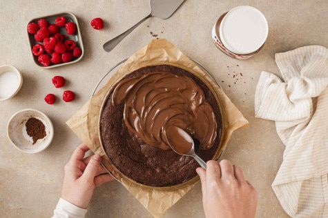 Draufsicht auf Hände, die nutella®-Glasur auf einen Schokoladenkuchen streichen, umgeben von Himbeeren und einem nutella®-Glas.