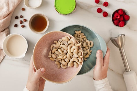 Draufsicht auf Hände, die eingeweichte Cashews für die Creme in eine andere Schüssel umfüllen.