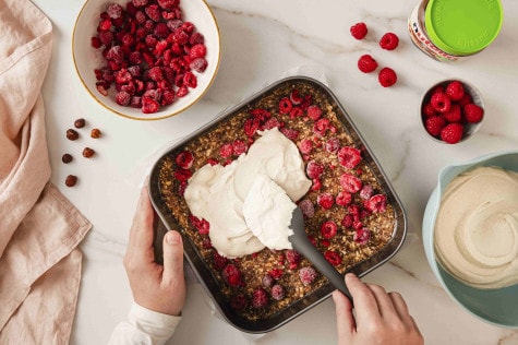 Draufsicht auf Hände, die Cashewcreme über eine Mischung aus Nüssen und Himbeeren in einer Backform verteilen.