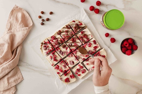 Draufsicht auf Hände, die nutella® auf Himbeerdessertquadrate träufeln. Haselnüsse und Himbeeren liegen daneben.