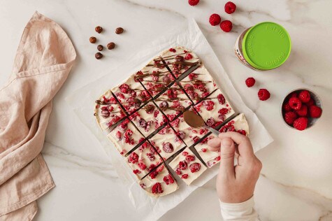 Draufsicht auf Hände, die nutella® auf Himbeerdessertquadrate träufeln. Haselnüsse und Himbeeren liegen daneben.