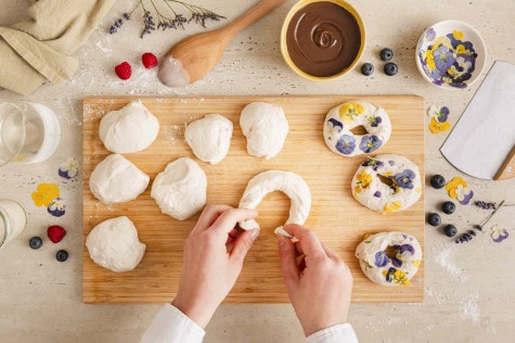 Geformte Bagel-Teiglinge mit essbaren Blüten werden auf Holzbrett von Hand geformt