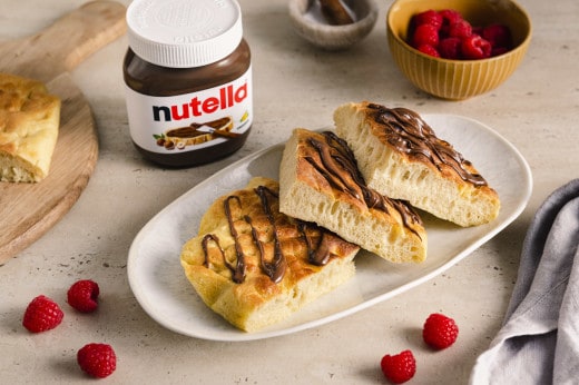 Luftige Focaccia-Stücke mit nutella®-Topping auf einem Teller, daneben Himbeeren und ein Glas nutella®