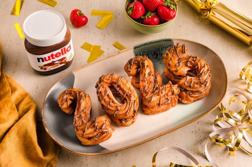 Nutella-Glas mit Messer, geschnittenem Brot und Zutaten im unscharfen Hintergrund.