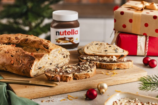 Ein frisch gebackener Weihnachtsstollen liegt auf einem Holzbrett. Ein Stück Stollen ist bestrichen mit nutella® und garniert mit Haselnüssen. 