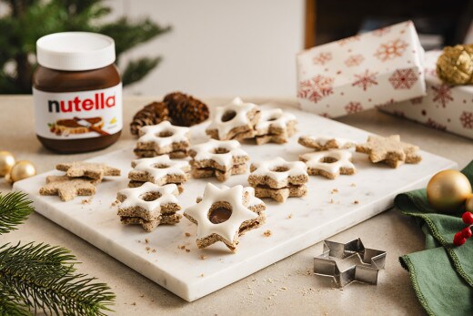 Mehrere frisch gebackene und glasierte Zimtsterne liegen auf einer weißen Marmorplatte. Neben der Marmorplatte steht ein Glas nutella®.