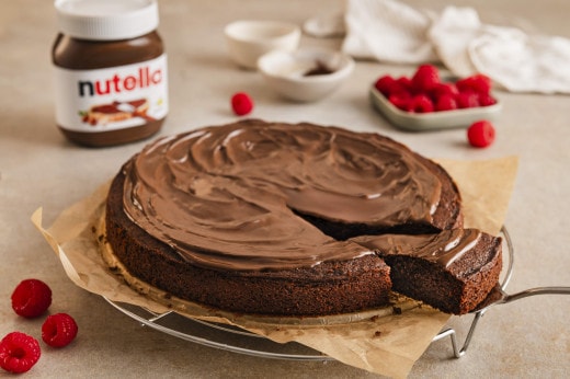 Ein Schokoladenkuchen mit nutella®-Glasur, ein Stück davon abgeschnitten und mit Himbeeren bestreut, daneben ein nutella®-Glas.