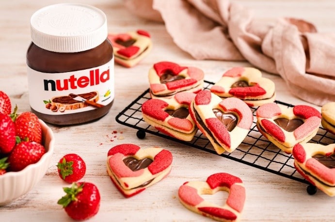 Herzförmige Cookies mit Nutella® und frischen Erdbeeren, serviert auf einem Gitter neben einem Glas Nutella®.