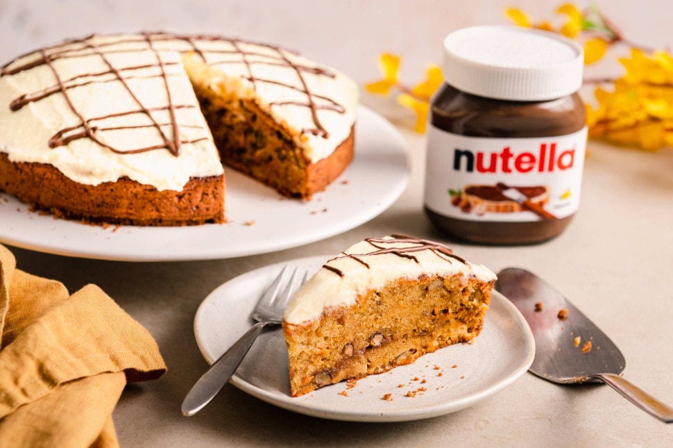 Ein Stück Karottenkuchen auf einem kleinen Teller, im Hintergrund der ganze Kuchen mit nutella®-Tröpfchen.