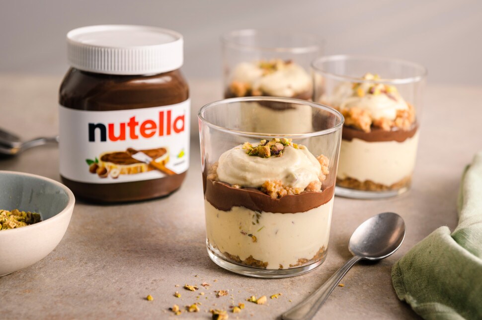 Pistazien-Cheesecake im Glas mit nutella®, geschichtet mit Keksboden, Creme und Topping, im Vordergrund ein Löffel und ein Glas nutella®
