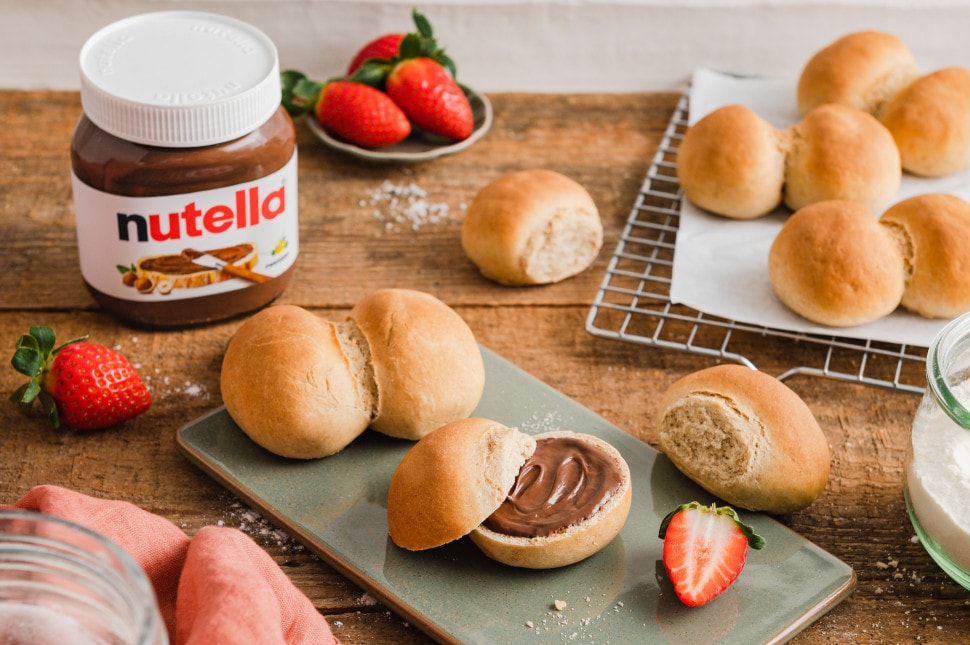 Picknickszene: Nutella-Glas, Kaffee und frische Backwaren auf Decke im Freien.